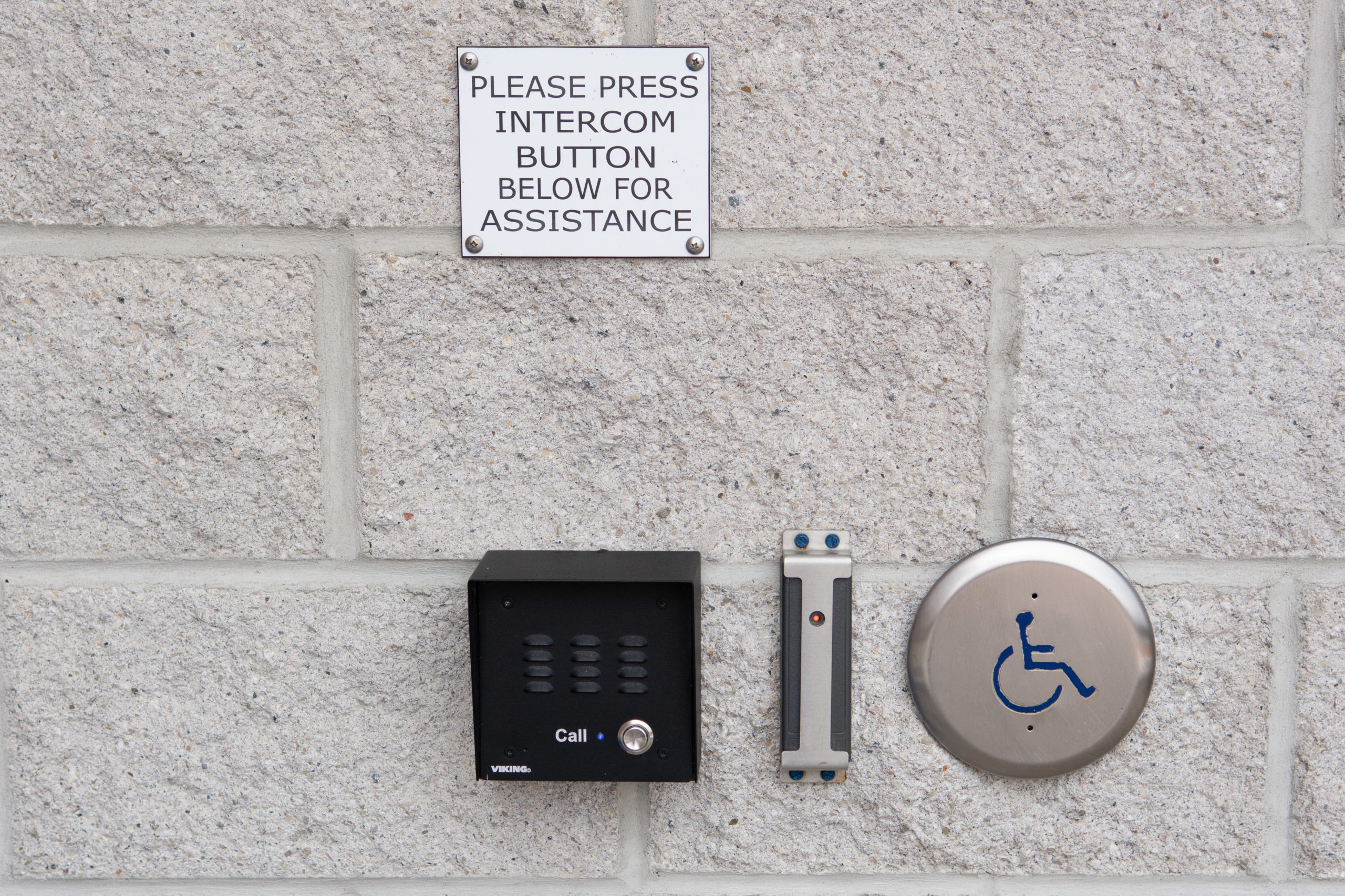 A black intercom box on a wall, beside an automatic door button with a sign above that reads "Please press intercom button below for assistance"