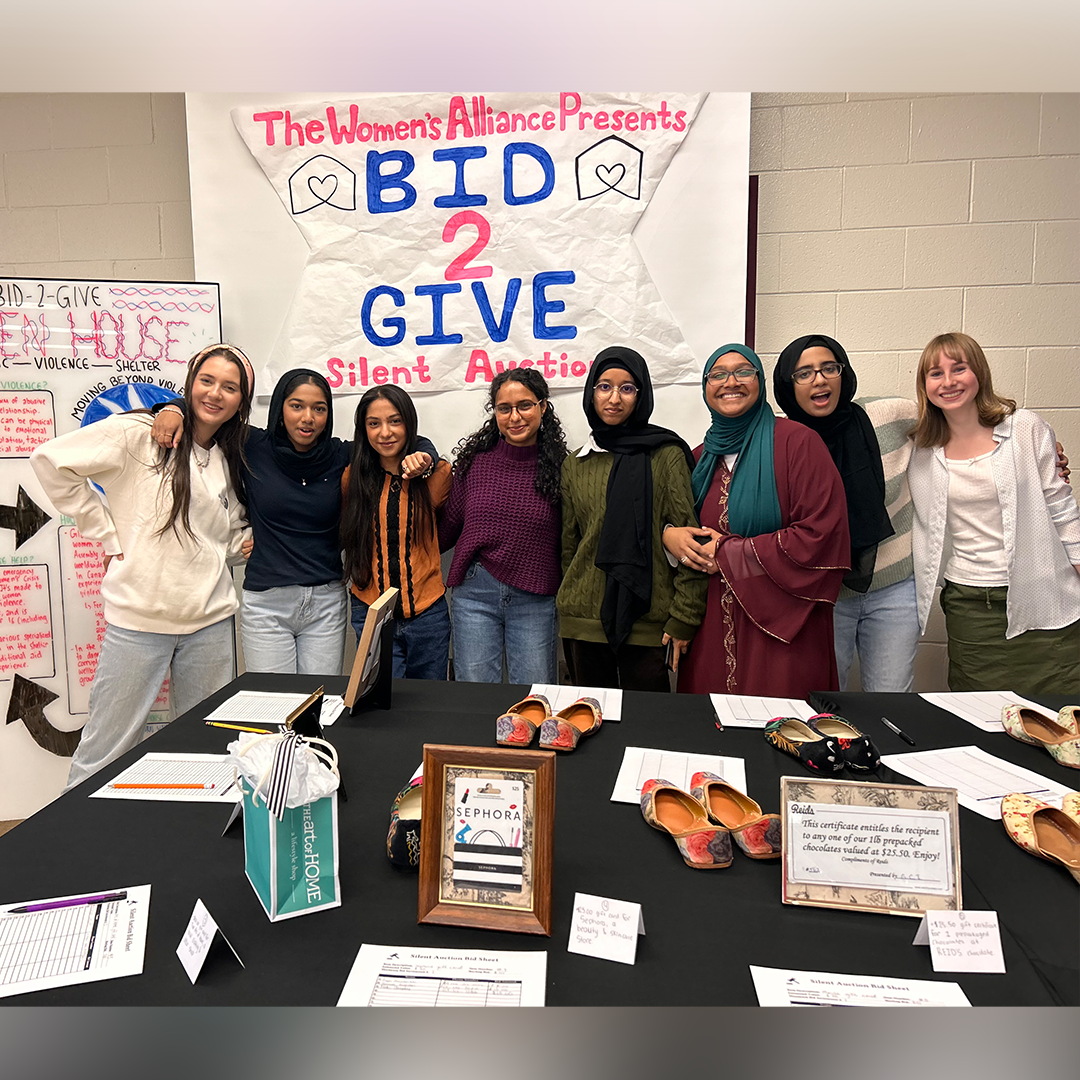 Students pose together in front of a banner that reads "The Women's Alliance Presents Bid 2 Give Silent Auction". 