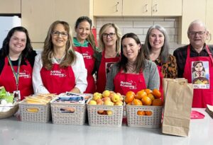 A group of seven people stand smiling behind a counter covered with food donation baskets. Each person is wearing a red apron with the "Nutrition 4 Learning" logo, and baskets are filled with fruit and other items.