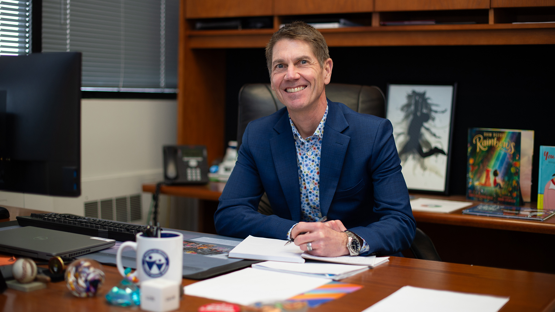 Scott Miller sits at his desk wearing a blue suit, pen in hand as he writes in a notebook, smiling towards the camera.