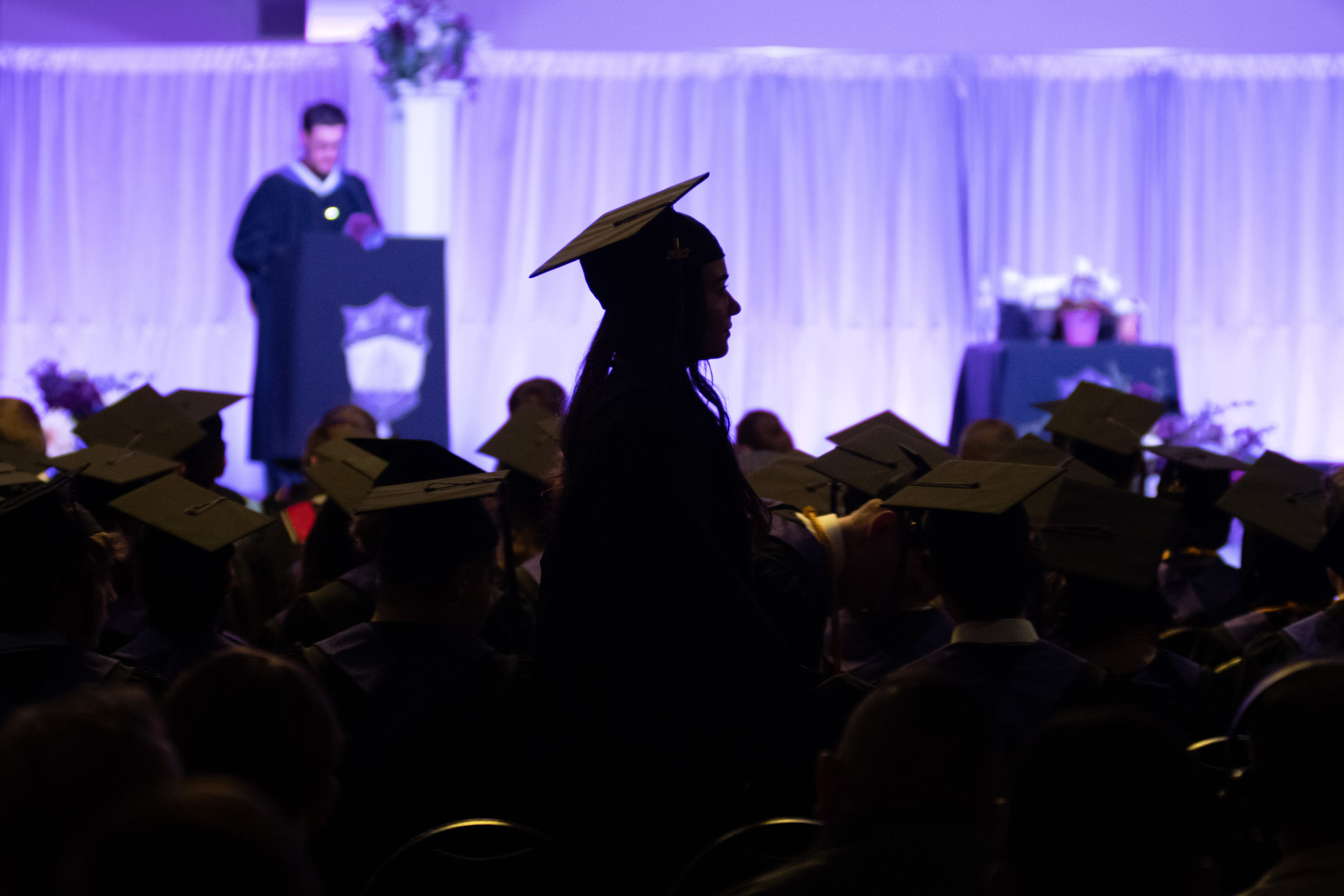 A student in a graduation cap is silhouetted against a purple-lit background as they walk to go and receive their diploma. 