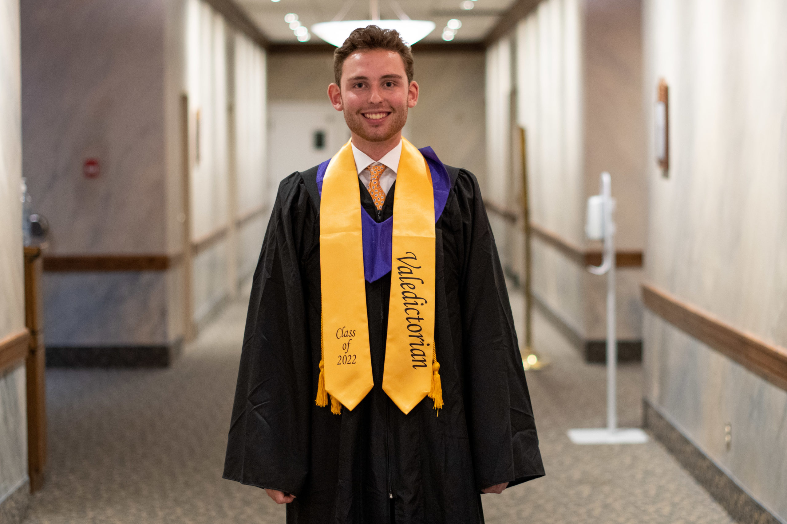 A student stands in the centre of frame, smiling towards camera wearing their graduation robes and valedictorian sash. 