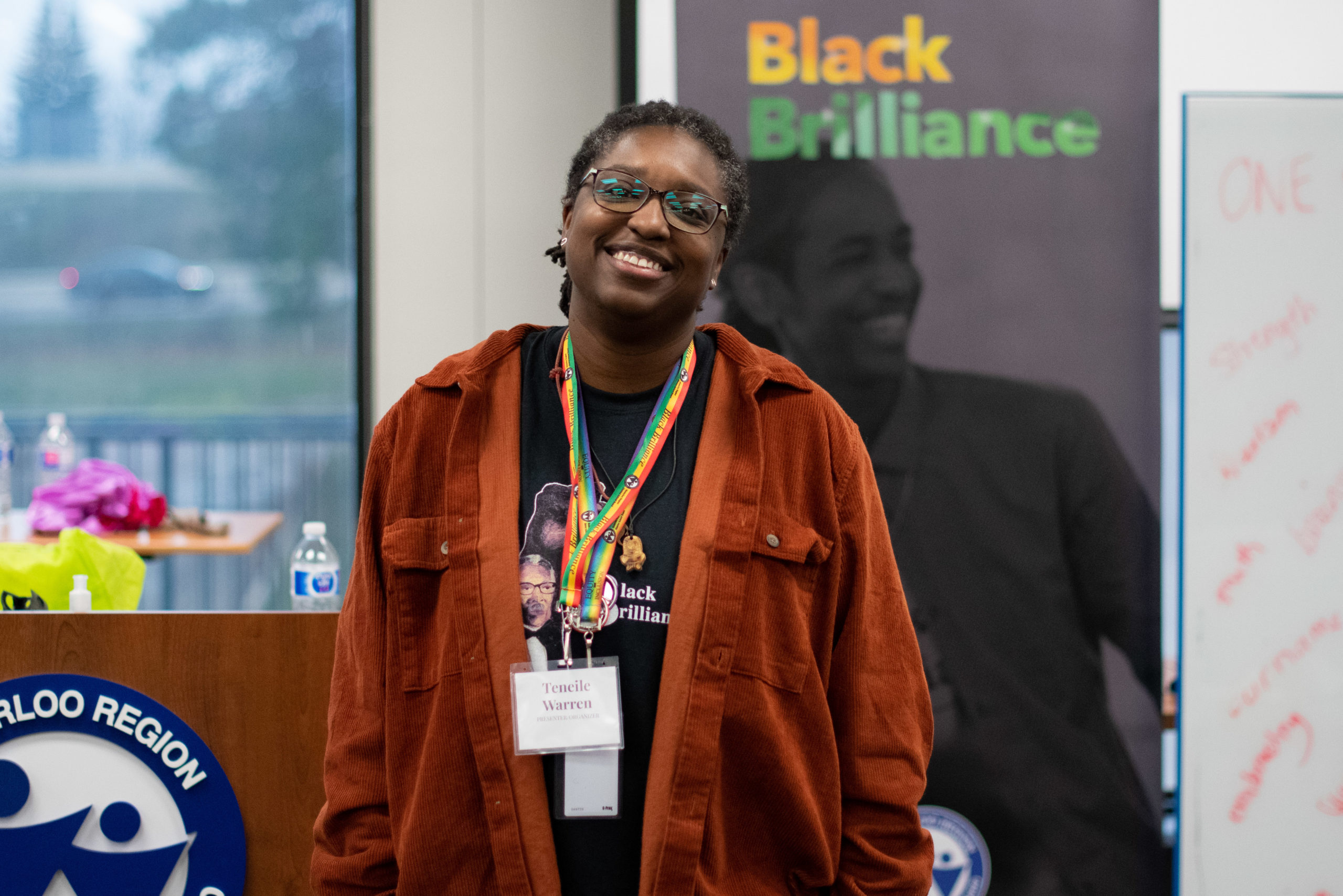 Teneile Warren looks towards the camera and smiles as they stand in front of a banner that reads "Black Brilliance". 